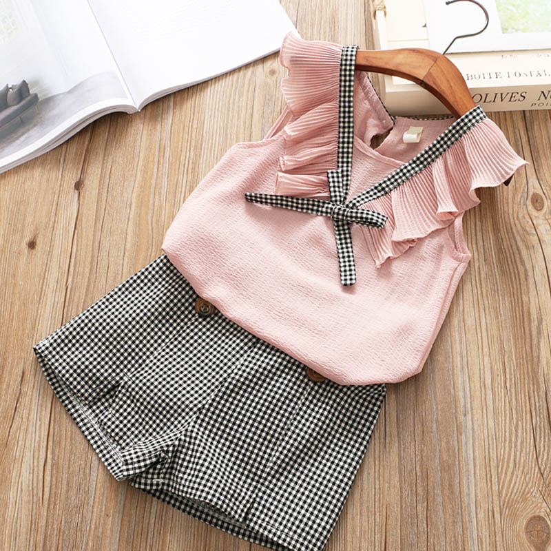 Girls short sleeve casual shirt and shorts set with pink ruffled top and checkered shorts on a wooden background.